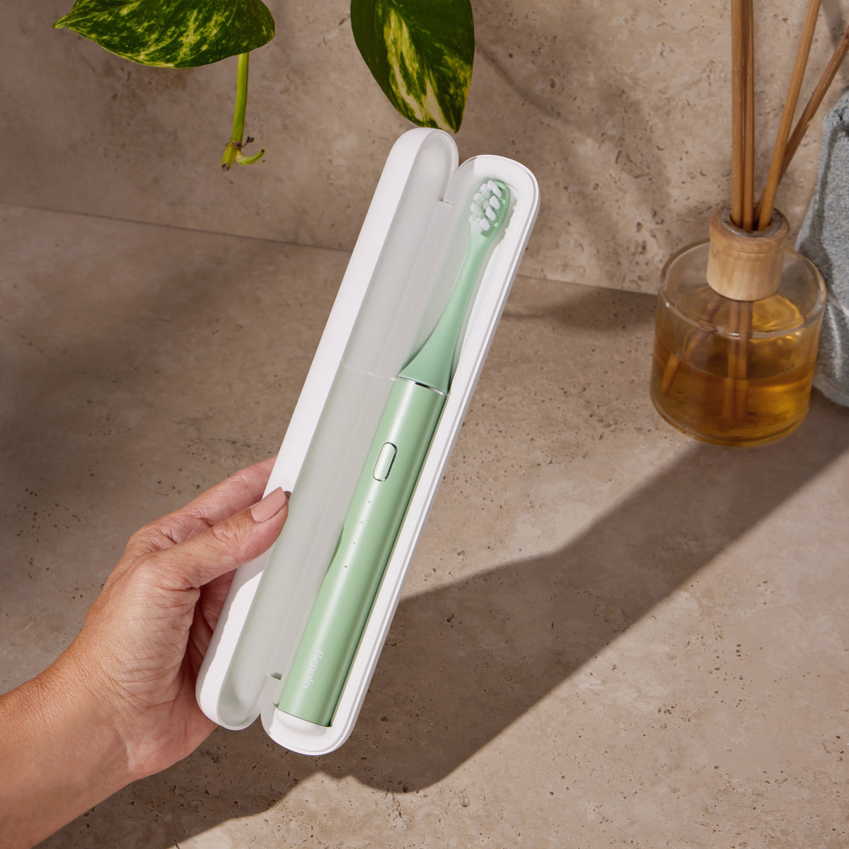 A hand holds an open white travel case containing a Bepulo green electric toothbrush against a beige stone wall and countertop, with a plant and reed diffuser in the background. #colour_pistachio green