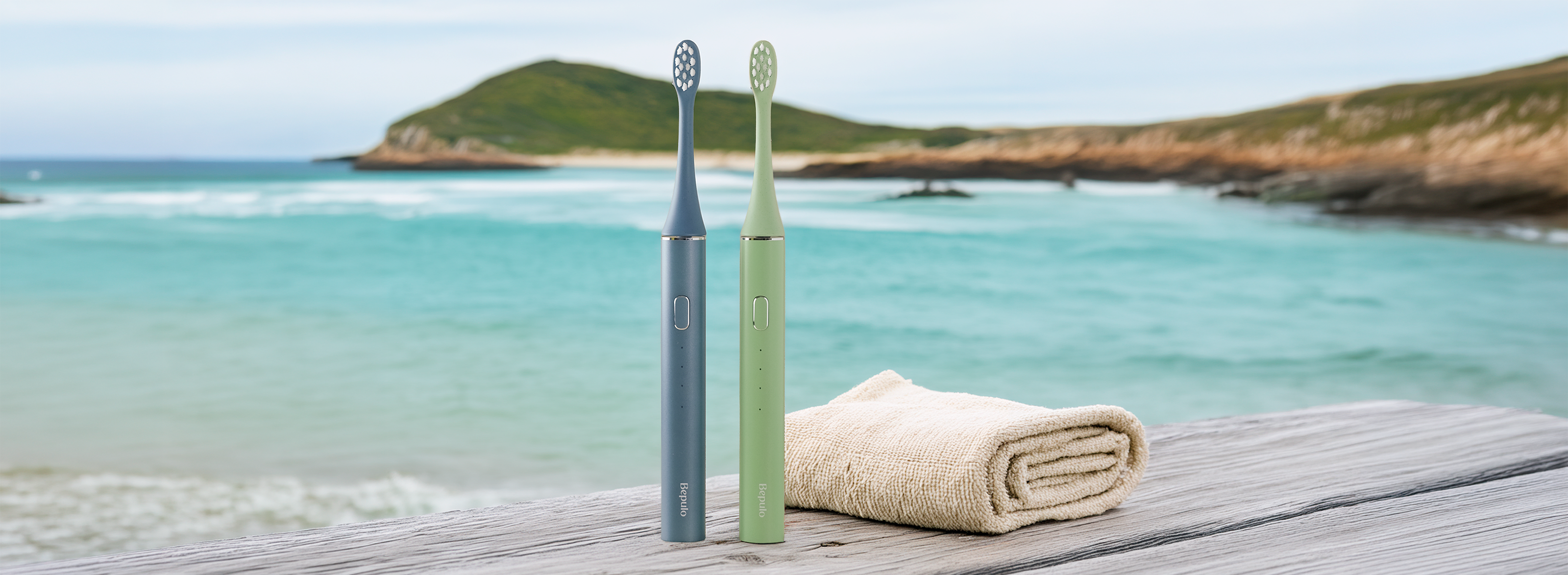 Two Bepulo - blue and green electric toothbrushes standing upright on a wooden deck with a rolled towel beside them, set against a scenic beach and ocean background.