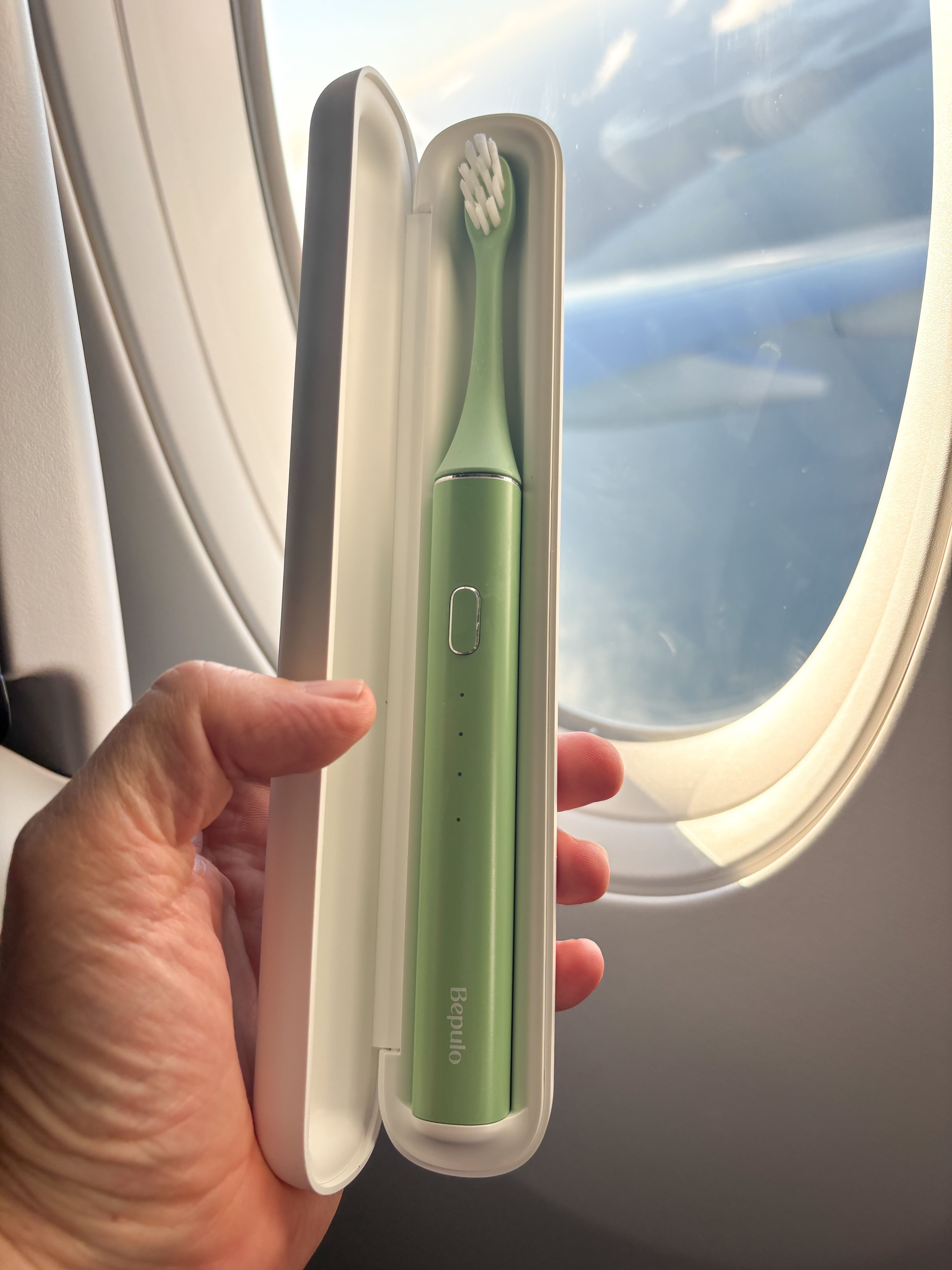 A person's hand holds an open white travel case containing a mint green electric toothbrush with white bristles, positioned next to an airplane window.