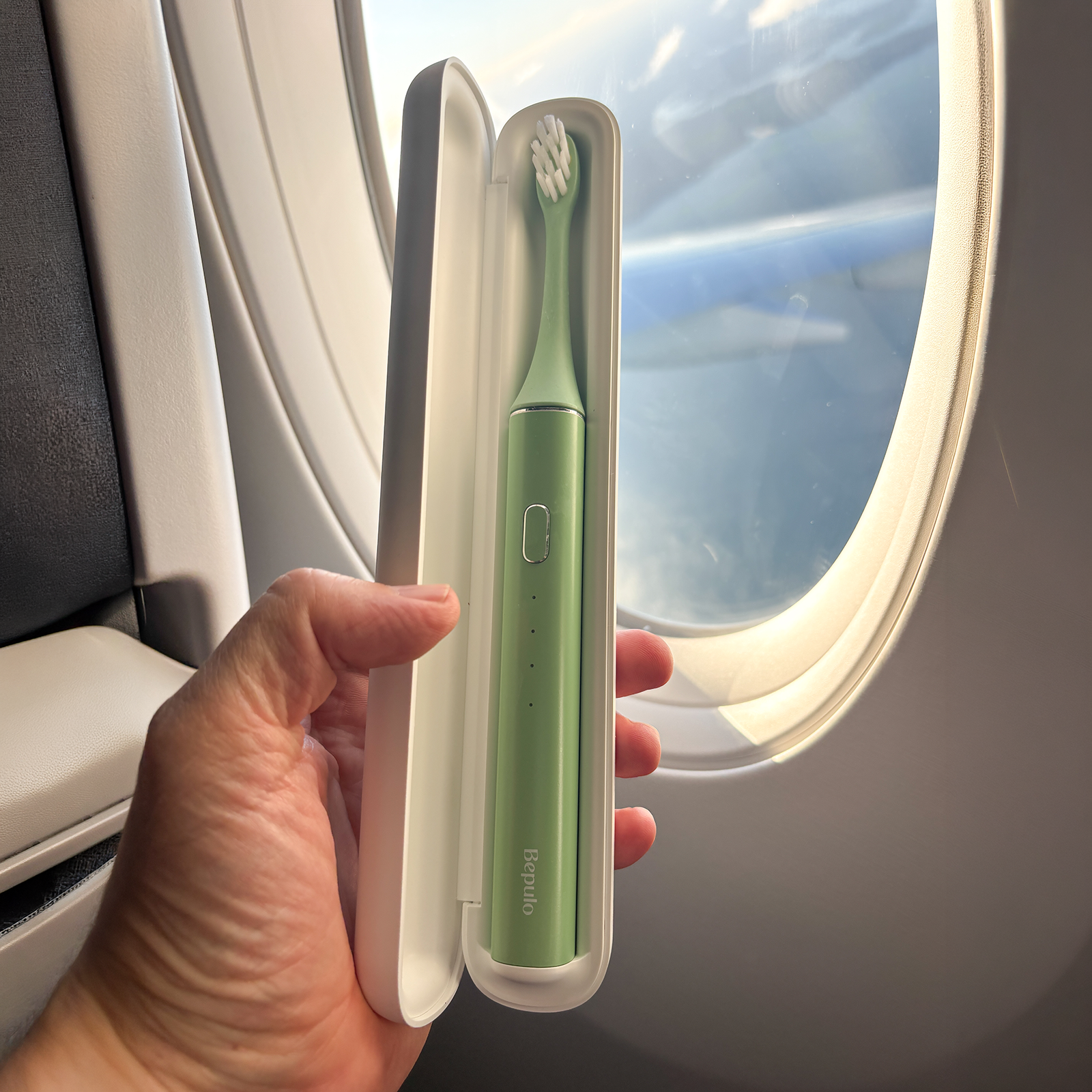 A hand holds an open white travel case containing a mint green electric toothbrush with white bristles, positioned next to an airplane window.