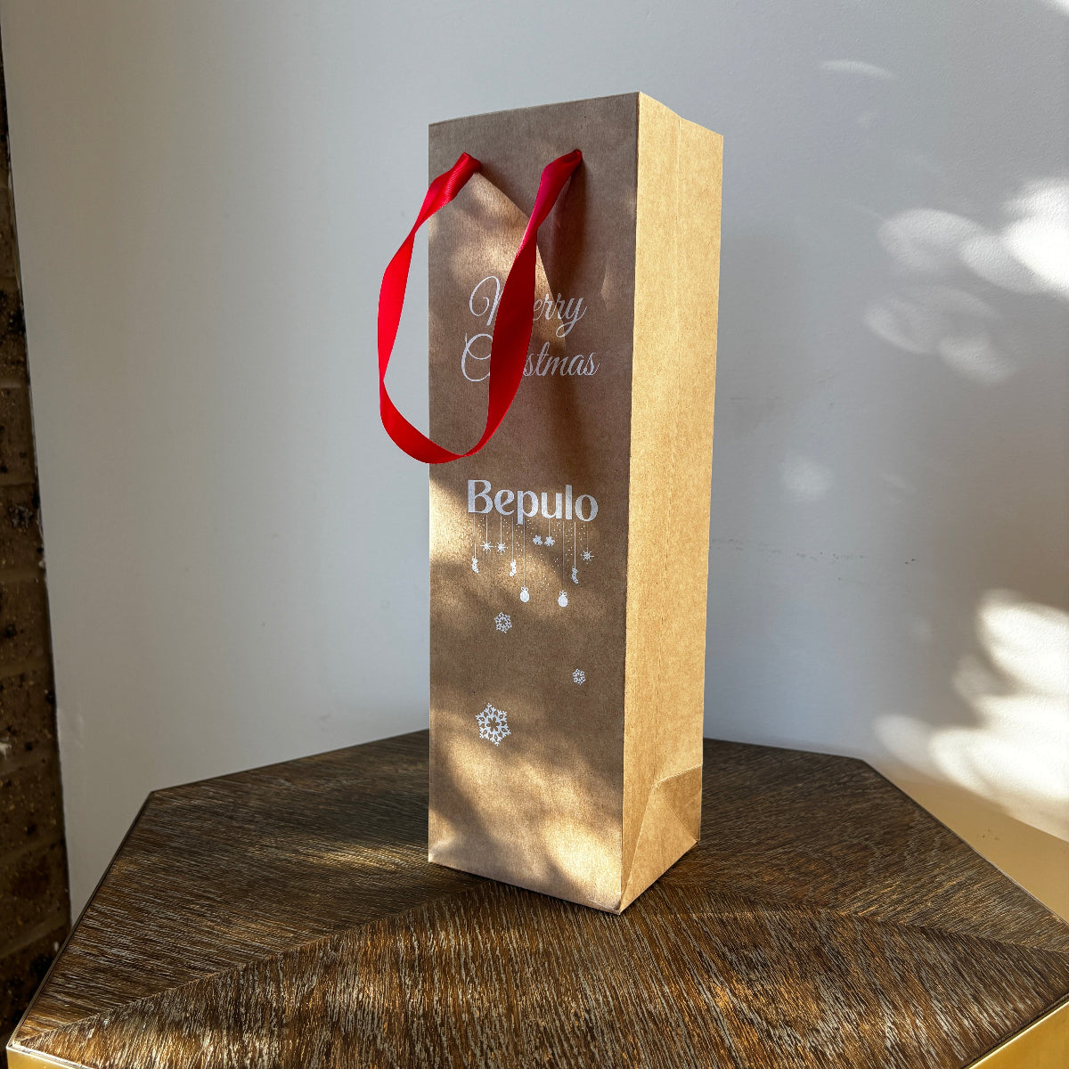 Bepulo white paper gift bag with red ribbon handles featuring "Merry Christmas" text on a wooden surface.