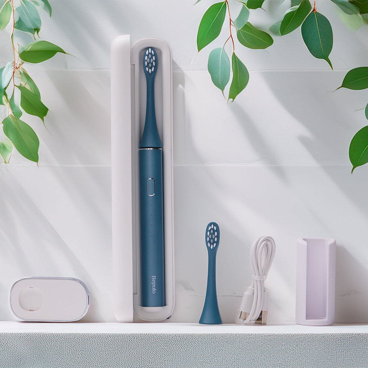 Bepulo Next Sonic Electric Toothbrush travel kit in Sapphire Blue with a white standing case, charging cable, wall mount, and replacement head on a clean spa bathroom shelf backdrop with green leaves. #colour_sapphire blue