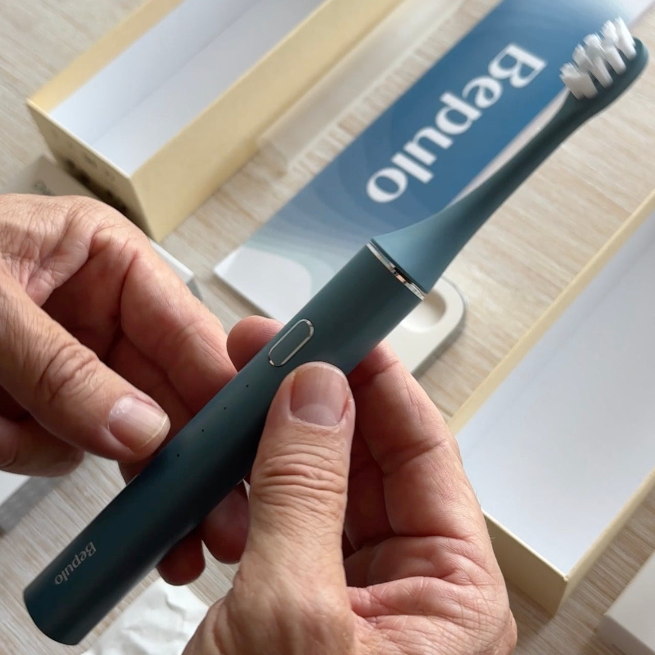 A person's hands holding a blue Bepulo electric toothbrush with white bristles, with its packaging box and blue booklet in the background.