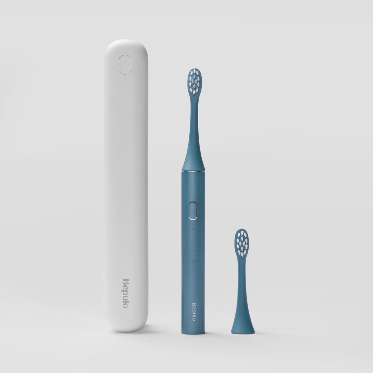 A Bepulo electric toothbrush in slate blue stands next to its matching blue replacement brush head and a tall white travel case on a light gray background. | #colour_sapphire blue
