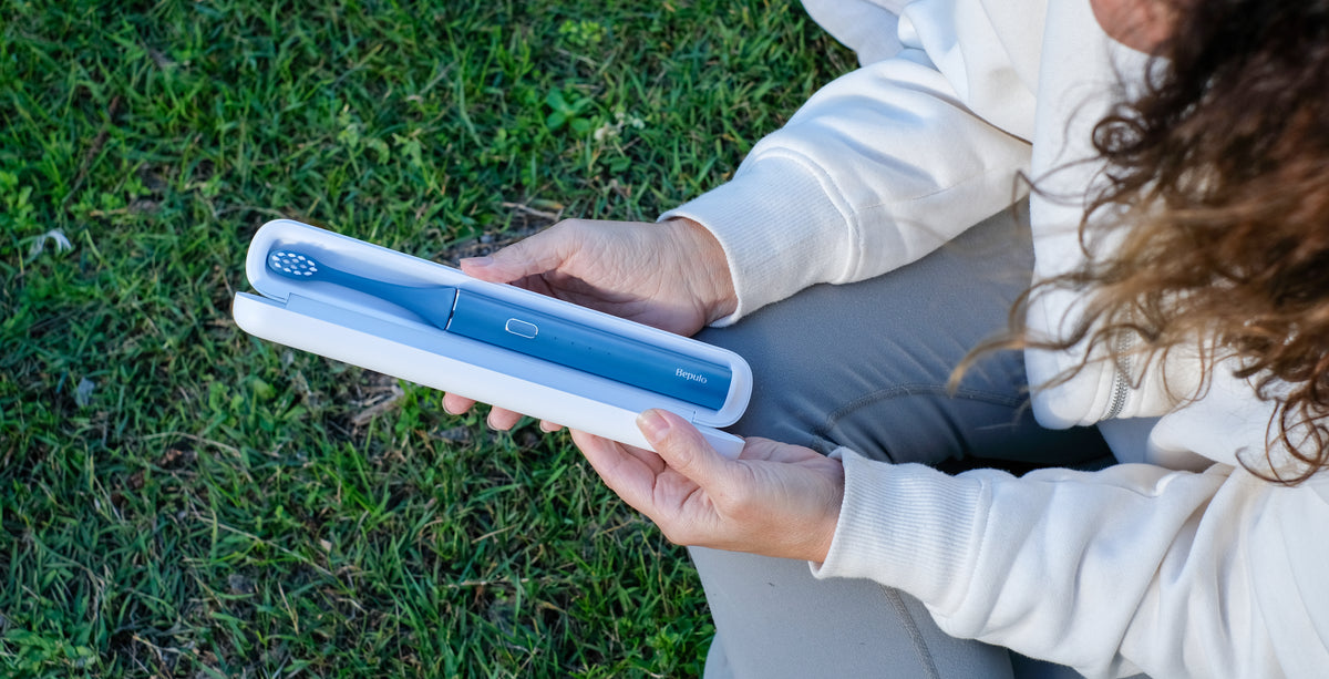 A person outdoors sitting on the grass holds a Blue Bepulo electric toothbrush in its white UVC Travel Case. 