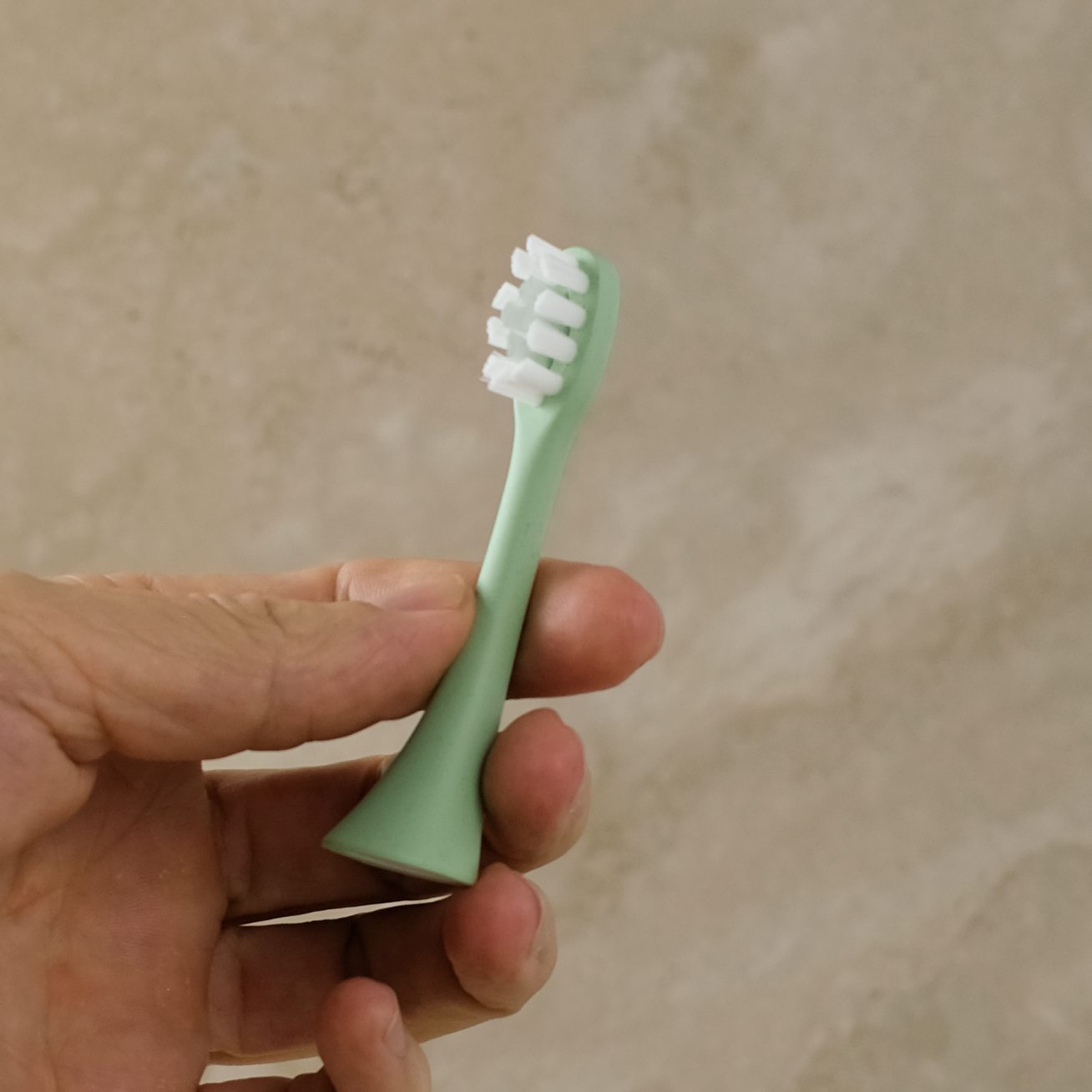 A hand holding a mint green replacement toothbrush head with white bristles against a textured beige background.