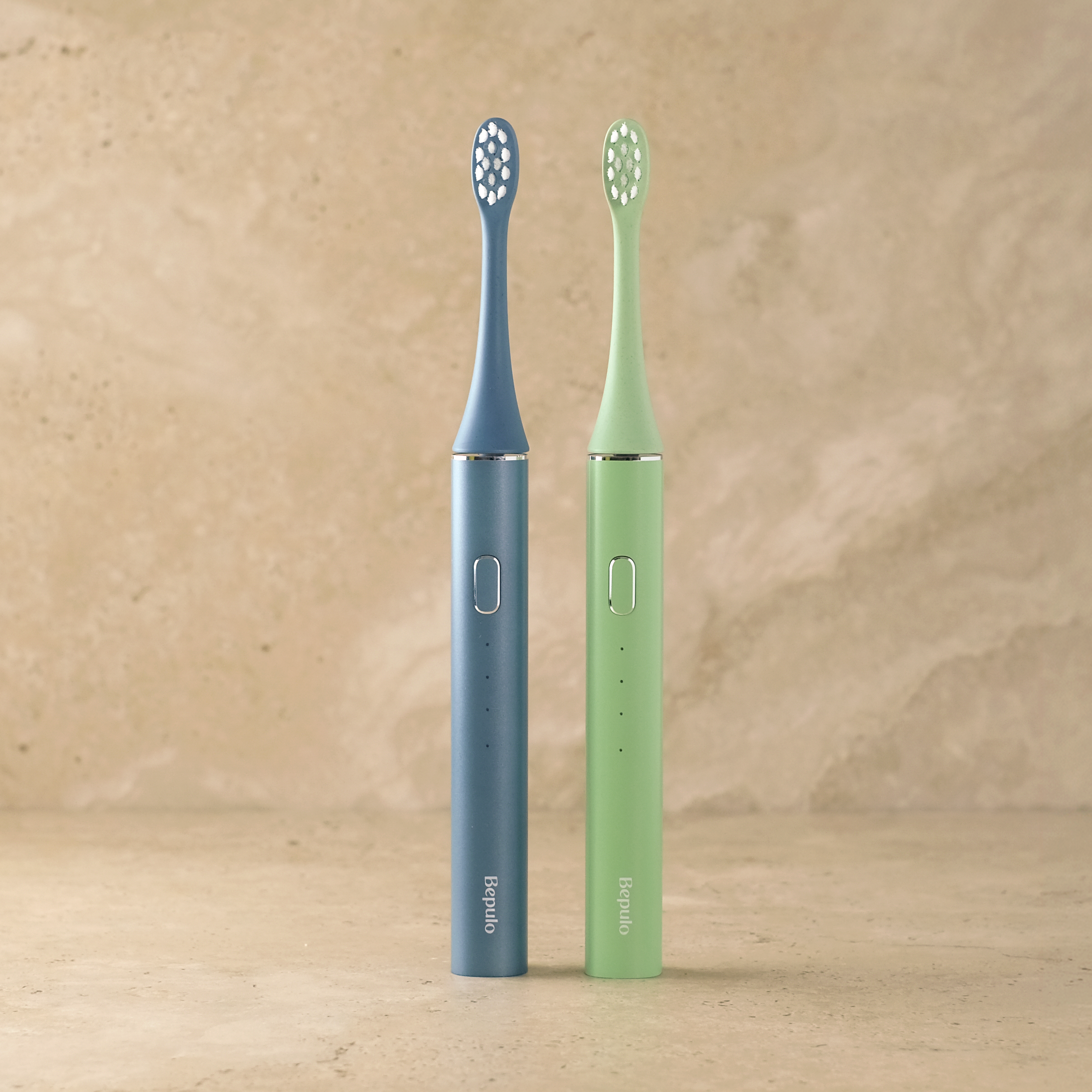Two Bepulo electric toothbrushes standing upright on a beige surface against a textured beige wall. One toothbrush is blue and the other is mint green, both with white bristles.