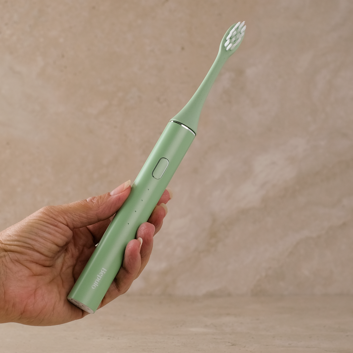 A hand holding a mint green electric toothbrush against a textured beige stone background.