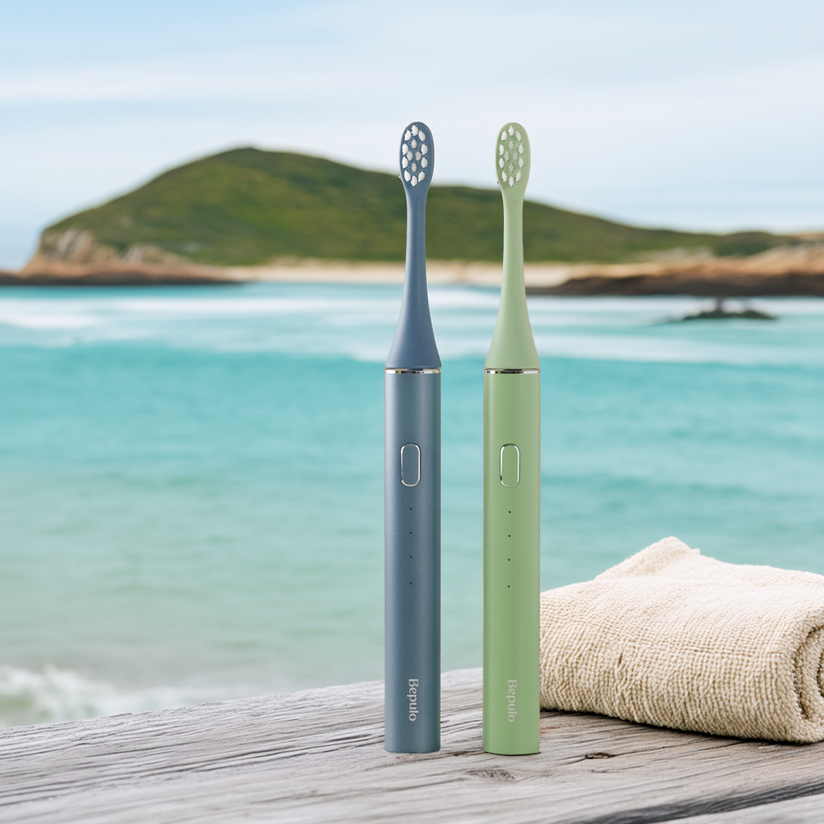 Two Bepulo electric toothbrushes standing upright on a wooden surface by the ocean, with a rolled beige towel beside them. One toothbrush is slate blue and the other is light green, both with gold-tone button accents.