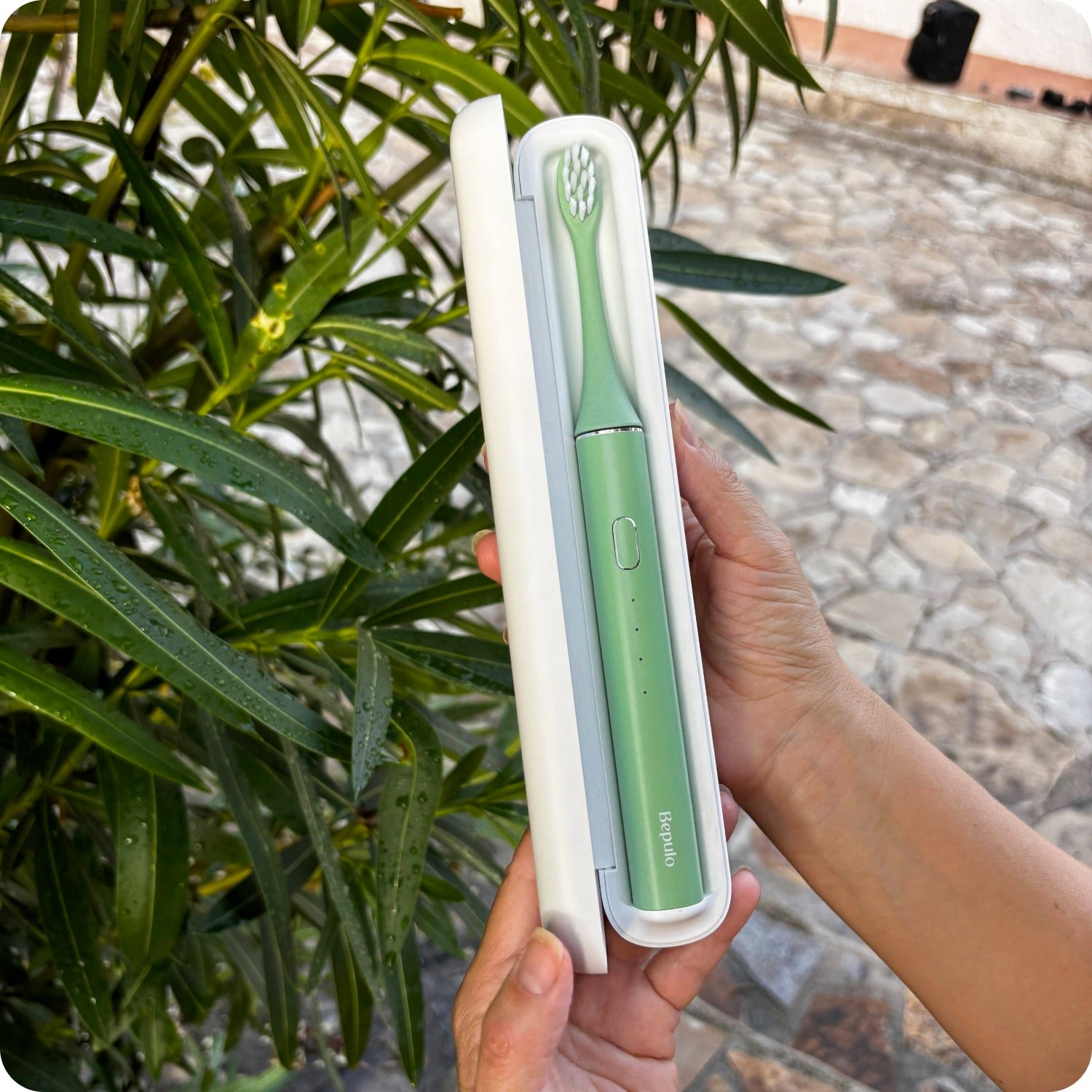 A person holds a compact electric toothbrush in a white travel case outdoors, with green leaves and a stone walkway in the background.