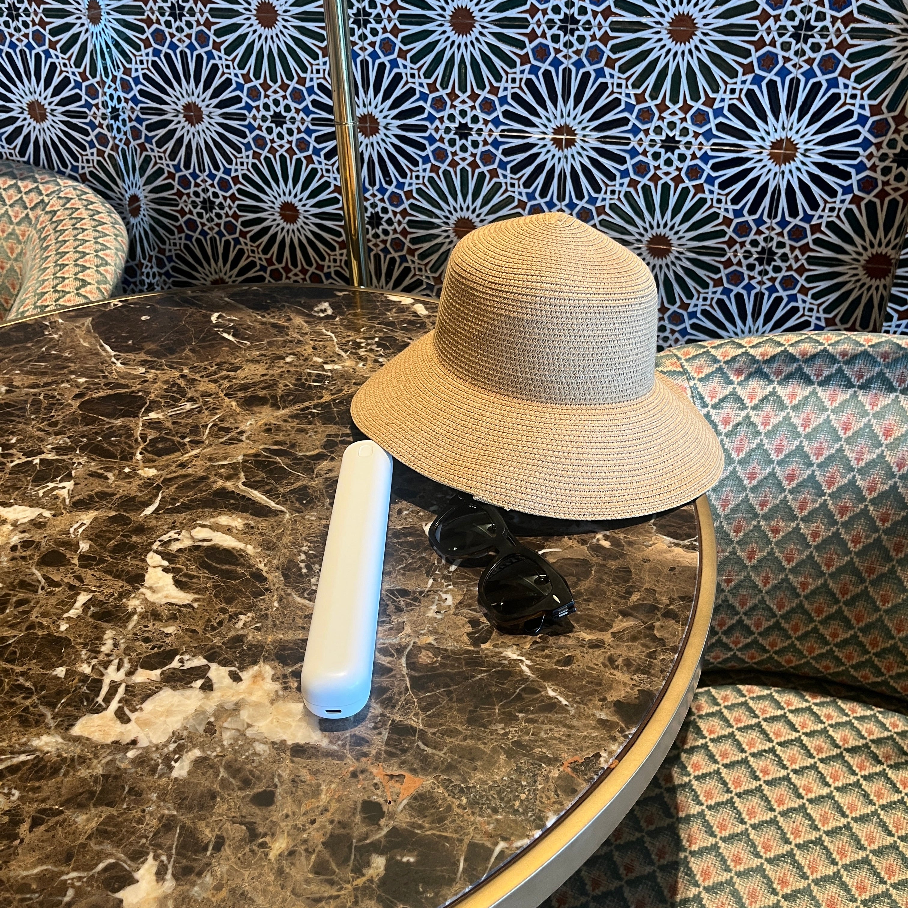 A light beige woven sun hat resting on a round dark marble table, with a white remote control and a black object beside it. The background features patterned blue, white, and black tiles on the left and textured green and beige upholstered seating on the right.