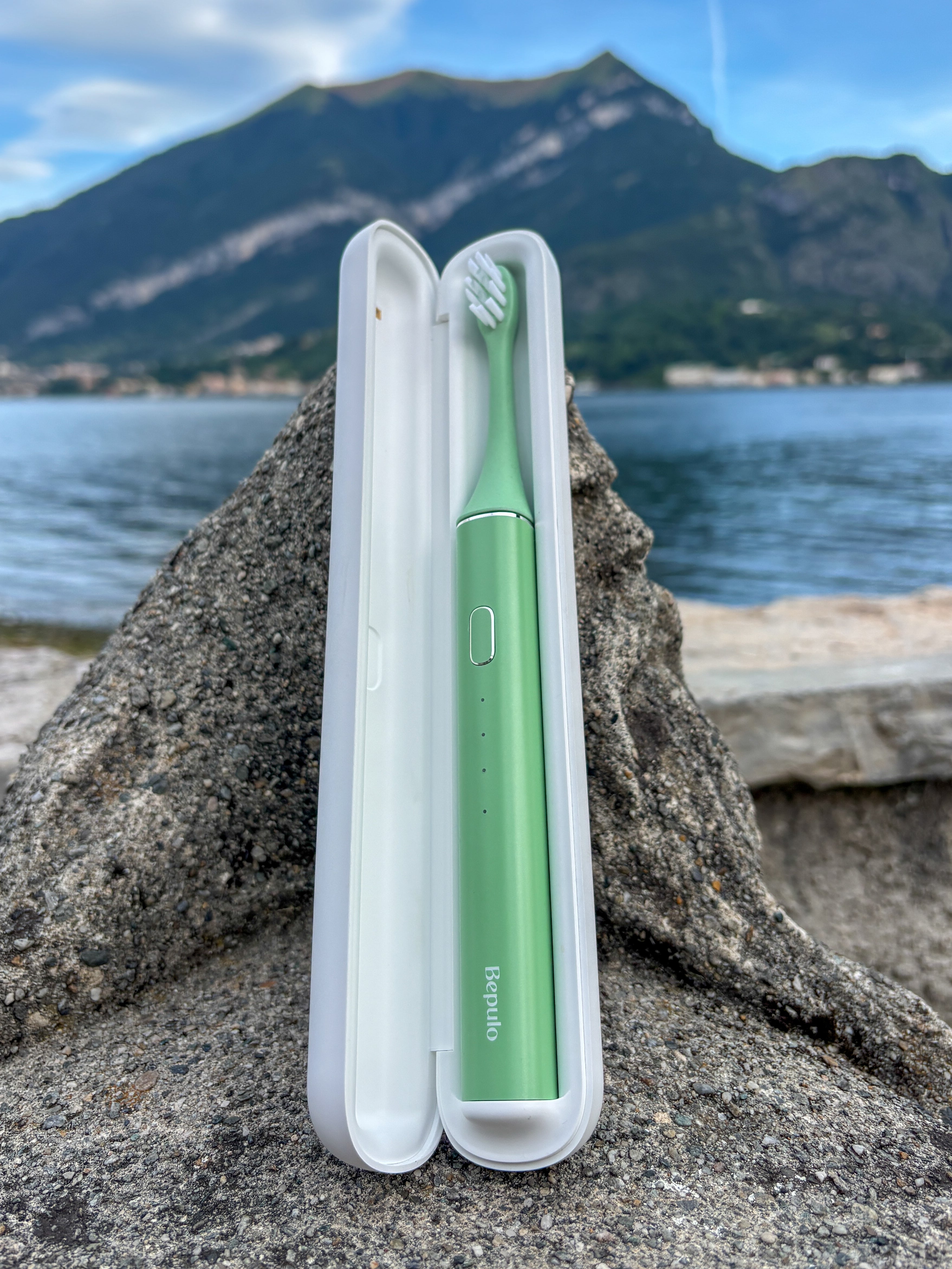 A mint green electric toothbrush in a white hard case, set on a large rock with a scenic lake and mountain background.